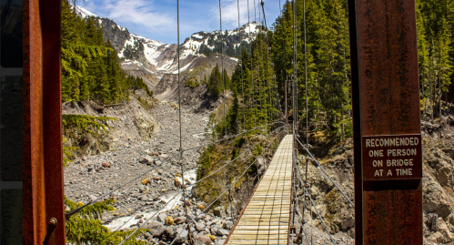 A suspension bridge over a rocky riverbed, surrounded by green trees and snow-capped mountains in the background.