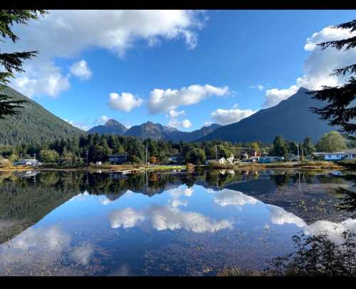 A serene landscape featuring a calm lake reflecting mountains and clouds, surrounded by trees and a small village.
