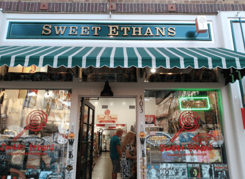 Exterior of "Sweet Ethans" candy store with a striped awning and colorful displays in the windows.