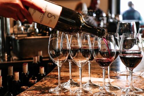 A hand pours red wine into several glasses lined up on a bar counter.