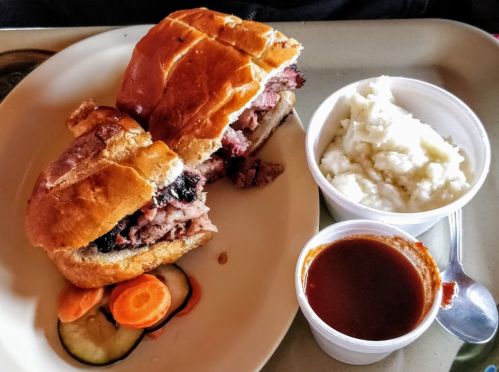 A plate with a sliced sandwich filled with meat, a side of mashed potatoes, and a small cup of sauce.