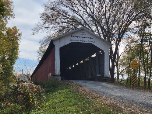 A red and white covered bridge surrounded by trees, with a sign indicating the bridge's name and year built.