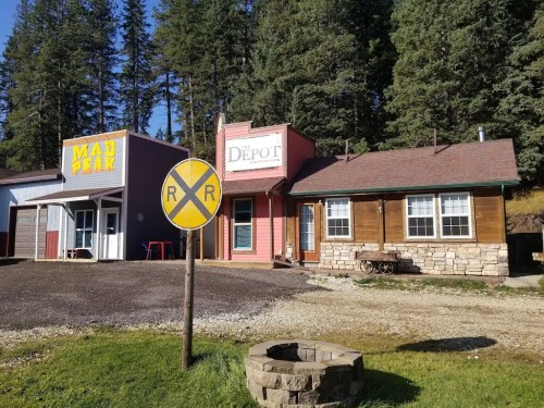 A quaint scene featuring two colorful buildings, "Mad Pear" and "Depot," beside a railroad crossing sign in a forested area.