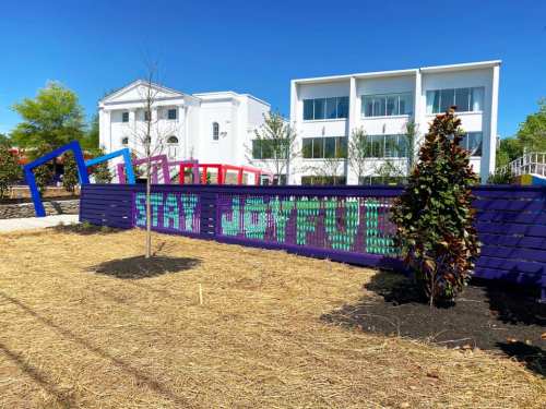 Colorful sign reading "Stay Joyful" in a park setting, with modern buildings and greenery in the background.