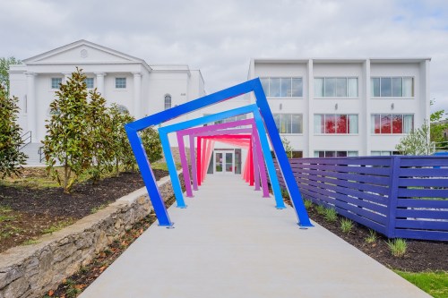 Colorful archway leading to a modern building, surrounded by greenery and a decorative fence.