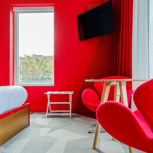 Bright red hotel room featuring a bed, a TV, and modern red chairs with a small table by the window.