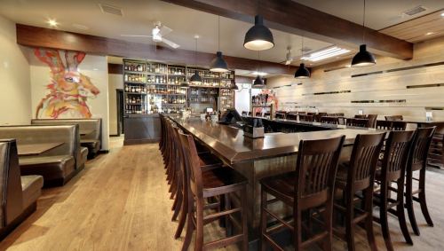A modern restaurant interior featuring a long bar, wooden seating, and artistic wall decor.