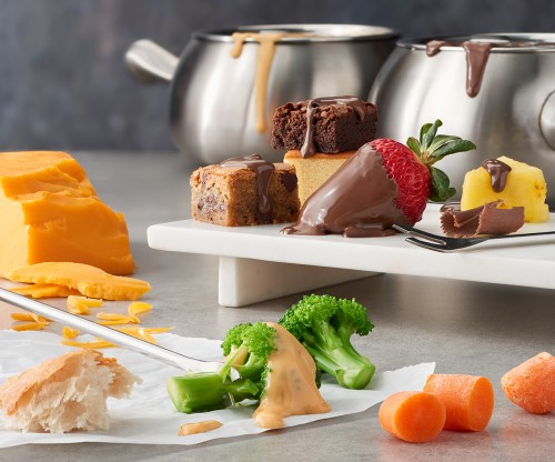 A platter with chocolate fondue, fruits, brownies, cheese, and vegetables, with dipping skewers and sauces.
