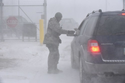 A person in winter gear hands something to a driver through a car window in a snowy, blizzard-like setting.