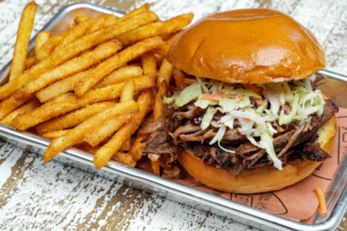A juicy pulled pork sandwich topped with coleslaw, served with a side of crispy fries on a metal tray.