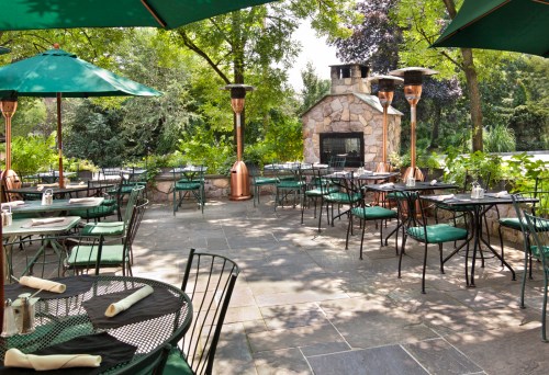Outdoor dining area with green umbrellas, tables, and a stone fireplace surrounded by lush greenery.
