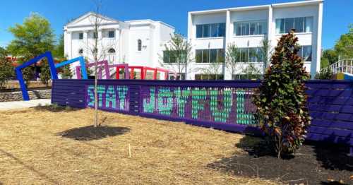 Colorful outdoor installation with the word "GRATITUDE" displayed on a fence, surrounded by greenery and modern buildings.