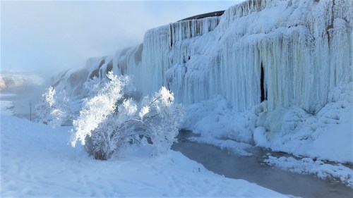 A frozen waterfall surrounded by snow-covered trees and mist, creating a serene winter landscape.