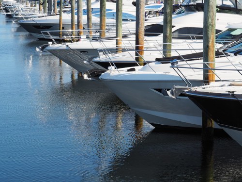 A row of boats docked at a marina, reflecting in calm water with wooden posts in the background.