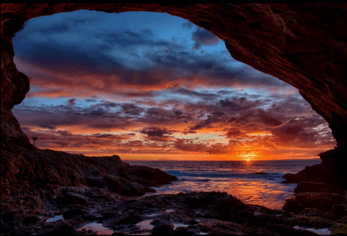 A stunning sunset viewed from a cave, with vibrant colors reflecting on the ocean and rocky shoreline.
