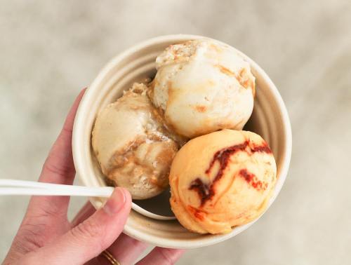 A hand holding a small bowl with three scoops of ice cream in different flavors.