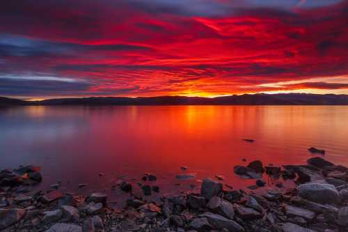 Vibrant sunset over a calm lake, with dramatic red and orange clouds reflecting on the water and rocky shoreline.