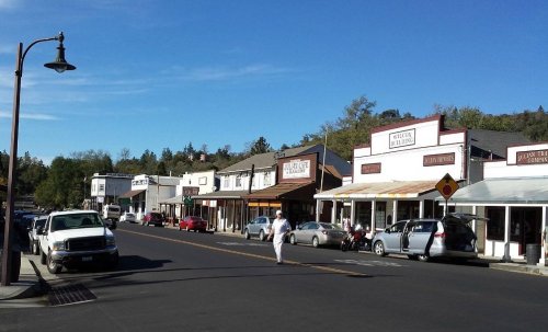 A quaint street lined with shops and parked cars under a clear blue sky. A person walks along the sidewalk.