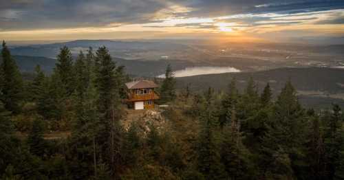 A scenic view of a cabin on a hillside, surrounded by trees, with a sunset over a valley and distant mountains.