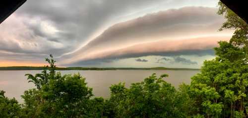 A panoramic view of a lake surrounded by trees, with dramatic, layered clouds in the sky at sunset.