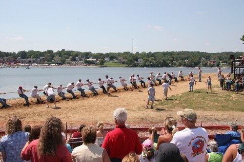 A tug-of-war competition by a lake, with teams pulling a rope while spectators watch from the shore.