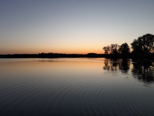 A serene lake at sunset, with calm waters reflecting the colorful sky and silhouettes of trees along the shore.