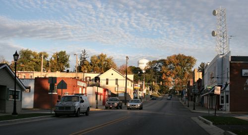 A quiet small town street with shops, trees, and a water tower under a cloudy sky. Cars are driving down the road.