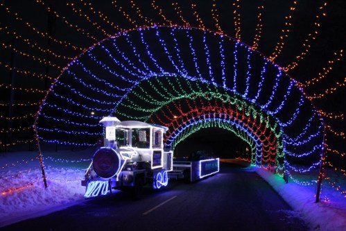 A brightly lit train surrounded by colorful holiday lights forming a tunnel on a snowy road at night.