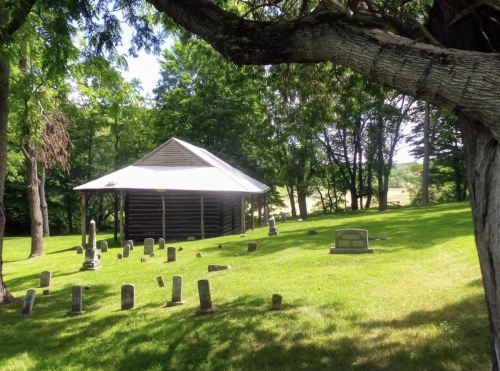 A peaceful cemetery with small gravestones, surrounded by trees and a rustic building under a bright sky.