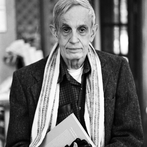 A man with gray hair and a scarf holds a book, wearing a blazer in a softly lit indoor setting.