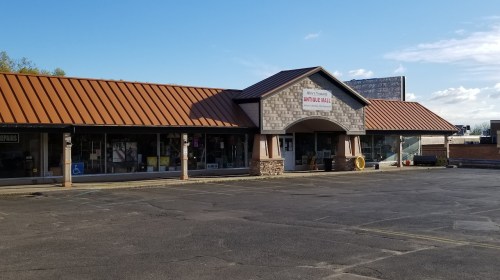 A single-story retail building with a brown metal roof and stone accents, featuring large windows and a parking lot.