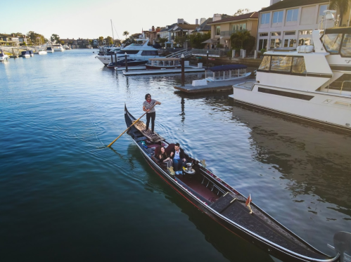 A gondola with people gliding through a calm marina, surrounded by boats and waterfront homes.