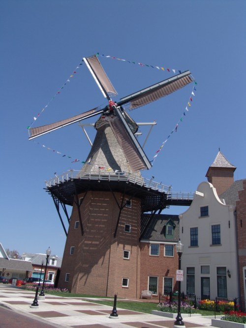 A large windmill with colorful flags, surrounded by buildings and flowers under a clear blue sky.