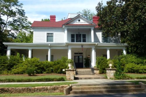 A large white house with a red roof, featuring a porch and surrounded by greenery and trees.
