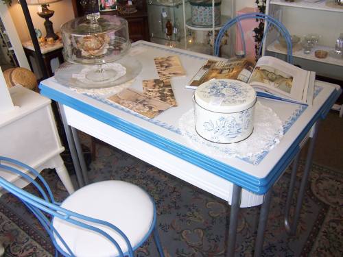 A vintage table with blue trim, a glass cake dome, old photographs, a magazine, and a decorative tin on a lace doily.