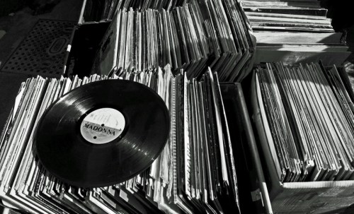 A close-up of a vinyl record resting on a stack of various records in black and white.