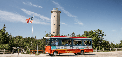 A colorful tour bus parked near a tall lighthouse and an American flag, surrounded by trees and blue sky.