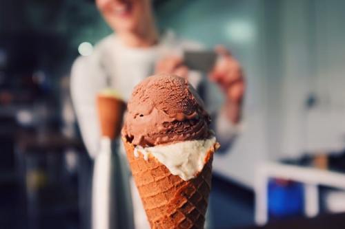 A close-up of an ice cream cone with chocolate and vanilla scoops, with a person taking a photo in the background.