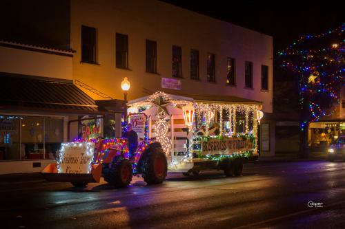 A decorated tractor pulling a festive float with lights, driving through a city street at night.