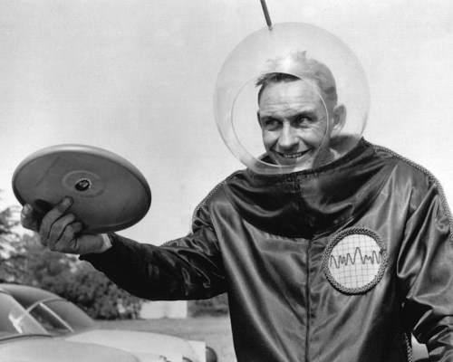 A man in a futuristic space suit and helmet smiles while holding a frisbee outdoors.