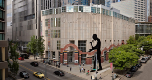 A modern building with large windows and a prominent sculpture outside, surrounded by trees and city streets.