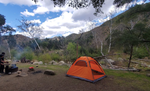 A vibrant orange tent set up in a scenic campsite surrounded by trees and mountains under a partly cloudy sky.