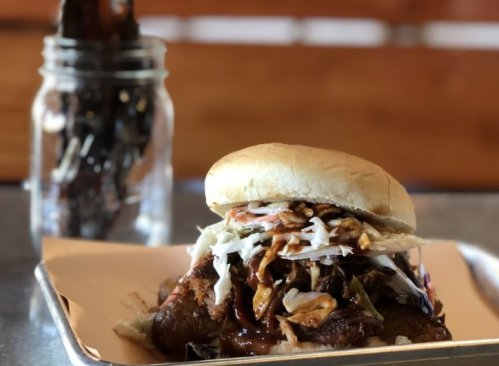 A close-up of a pulled pork sandwich topped with coleslaw, served on a tray with a jar of pickles in the background.