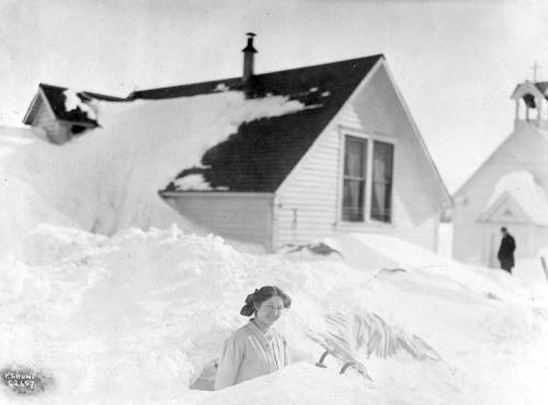 A woman stands in deep snow near a house, with snow piled high on the roof and a church in the background.