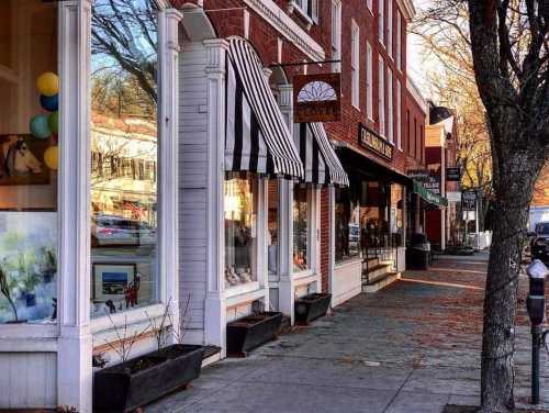 A charming street scene featuring brick buildings with striped awnings, shops, and autumn leaves on the sidewalk.