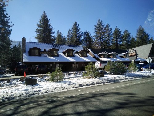 A rustic lodge with a snow-covered roof, surrounded by pine trees and parked cars along a clear road.