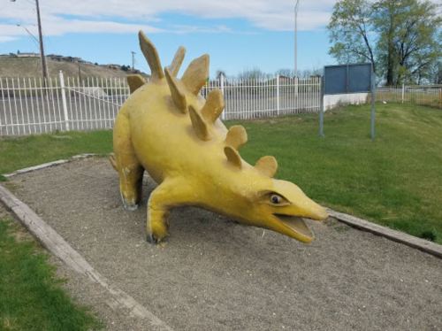 A large yellow dinosaur sculpture with spines, positioned in a grassy area near a fence.