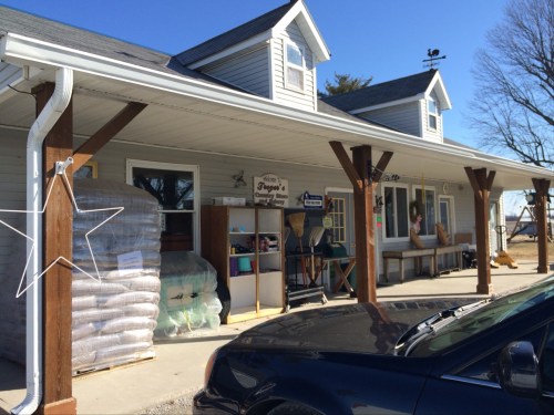 A small store with a porch, stacked bags outside, and a car parked in front on a sunny day.