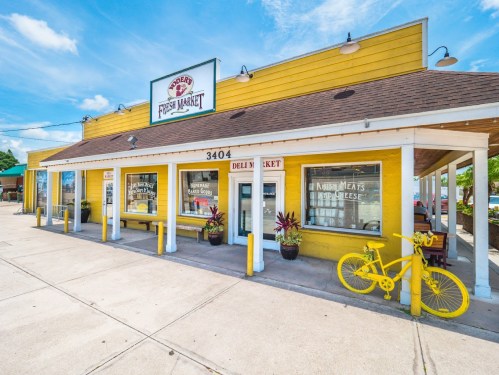 A bright yellow building with a sign reading "Fresh Market," featuring a bicycle and plants outside.