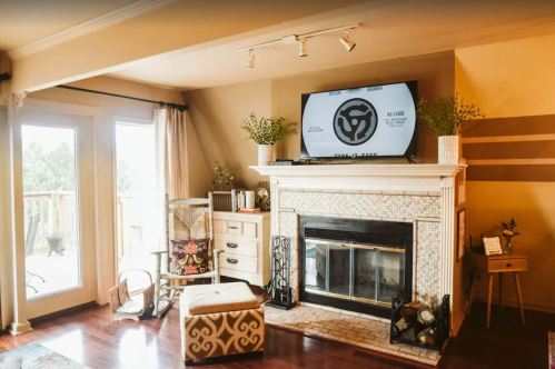 Cozy living room with a fireplace, TV above, and large windows. Decor includes plants and stylish furniture.